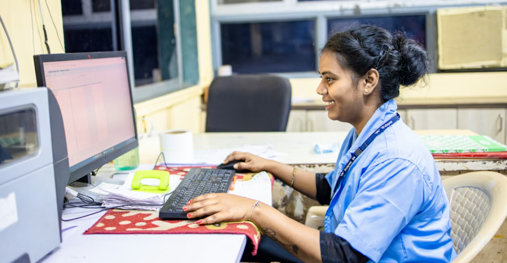 Een vrouw in een blauw uniform zit aan een bureau, werkt aan een desktopcomputer en glimlacht in een kantooromgeving, volgens Europese richtlijnen binnen de zorgsector.